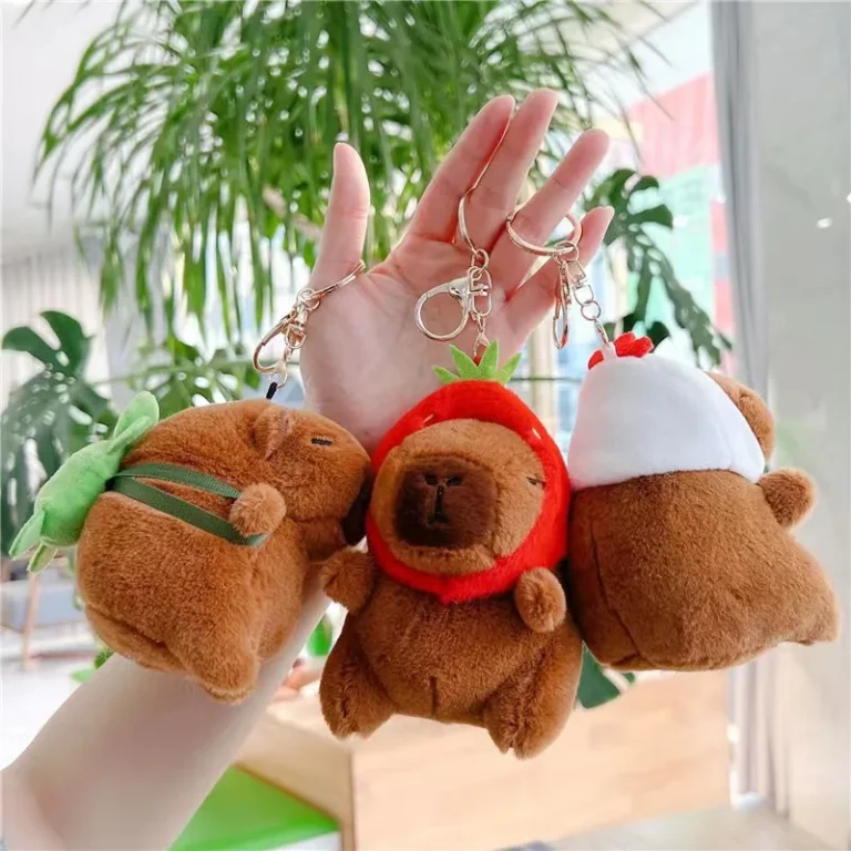 Three capybara plush keyrings held up in a hand in front of indoor plants