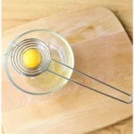 Spiral stainless steel egg separator over a glass bowl on a chopping board